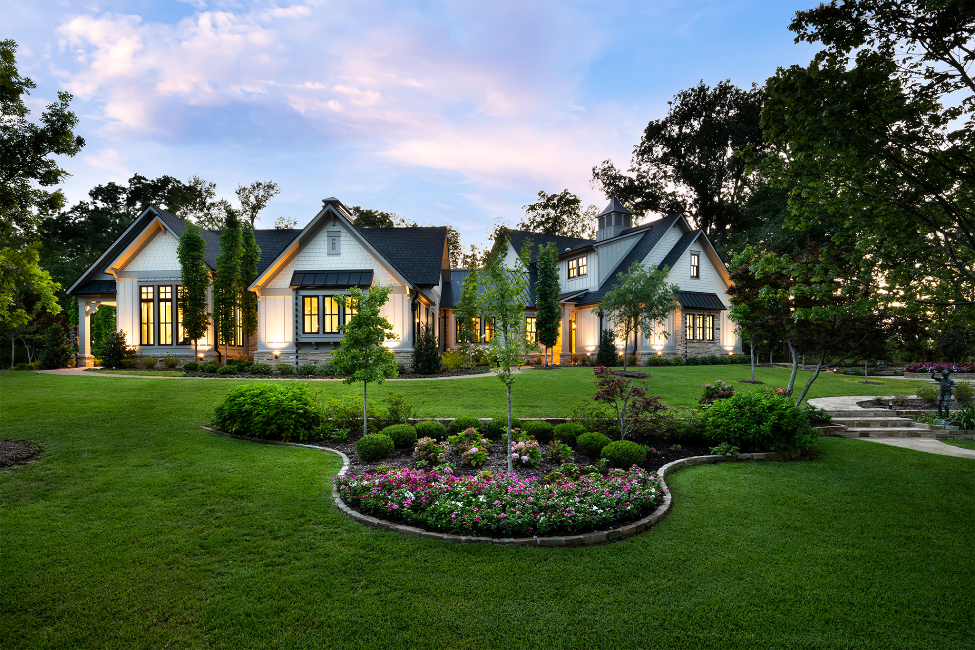 A modern suburban house with large windows, manicured lawn, flower beds, and surrounding trees at sunset.