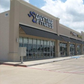 Exterior view of an Anytime Fitness gym in a strip mall with empty parking spaces in front.