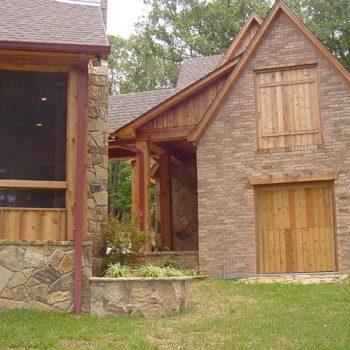 Brick and stone house with wooden accents, a covered window, and a wooden garage door, surrounded by grass and trees.