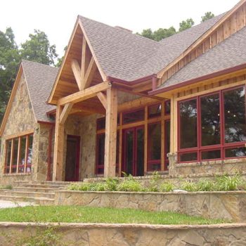 Stone and wood house with large windows, gabled roof, and landscaped yard surrounded by trees.