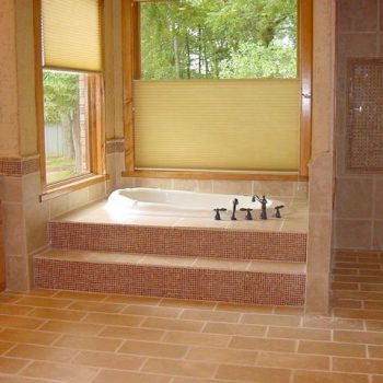 Large bathroom with tan tile flooring, built-in bathtub on tiled platform, and large windows with shades overlooking trees.