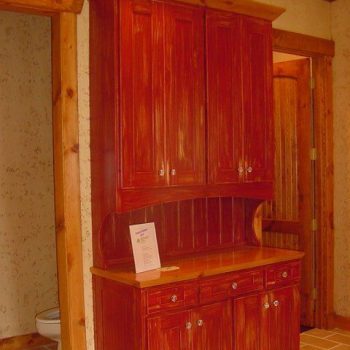 Red wooden cabinet with drawers and shelves stands against a wall near a bathroom door; a paper sits on the counter.