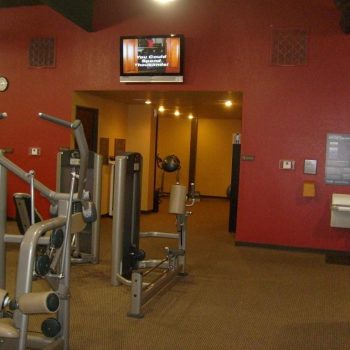Gym interior with exercise machines, red and tan walls, a mounted TV, water fountains, and informational posters.