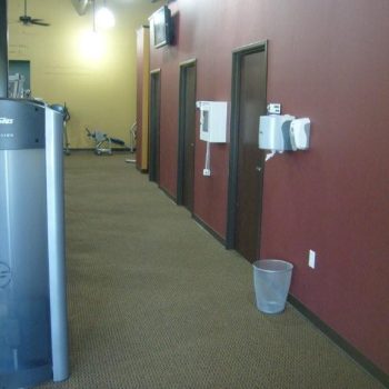 A gym hallway with maroon walls, brown doors, wall-mounted equipment, and a trash bin on a carpeted floor.
