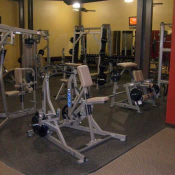 A gym room with multiple weight machines, benches, and mirrors, all set on a rubber mat floor.