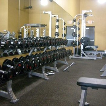 A gym weight area with racks of dumbbells, benches, weight machines, and mirrors on the wall.