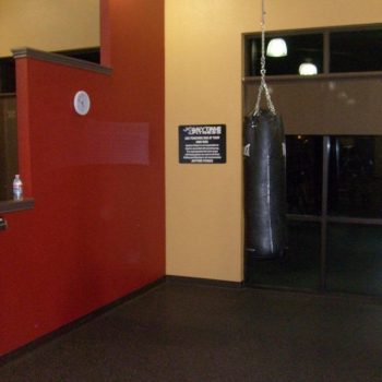 A black punching bag hangs from the ceiling in a gym corner with red and yellow walls and a window.