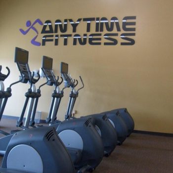 Row of elliptical machines in a gym with "Anytime Fitness" logo on the yellow wall in the background.
