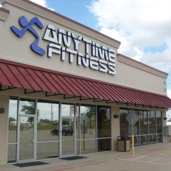 Exterior view of an Anytime Fitness gym with large windows and a red awning in a parking lot.