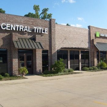 A brick building with signs for Central Title and H&R Block, featuring large windows and shaded entrances.