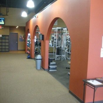 A gym area with red-orange archways, exercise equipment, carpeted floor, and a trash can near the entrance.