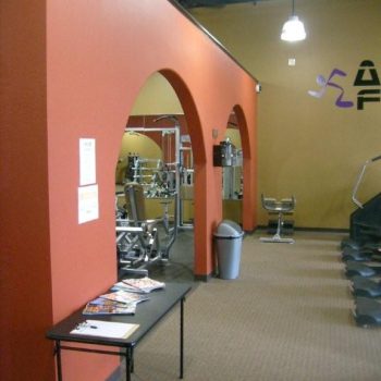 Fitness center interior with orange arches, gym equipment, a trash can, and a table with magazines in the foreground.