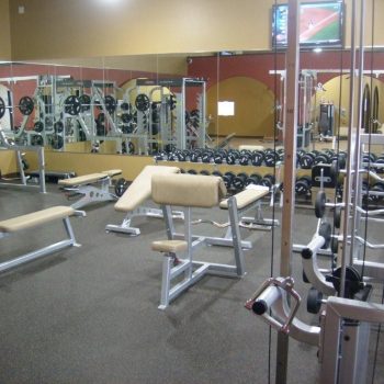 A gym room with weight benches, free weights, weightlifting machines, mirrors, and a TV on the wall.