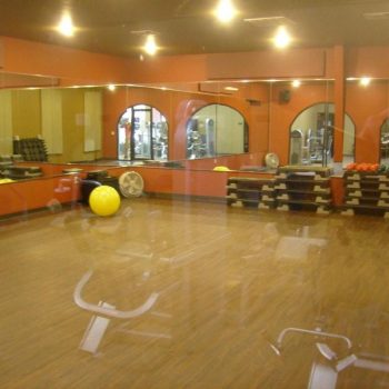 Empty fitness studio with mirrored walls, exercise balls, stacked steps, and view of gym equipment through glass windows.
