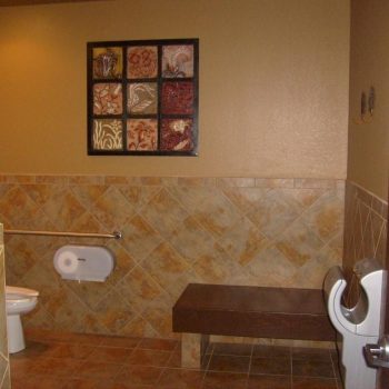 Accessible restroom with a toilet, wall-mounted bench, urinal, tiled walls and floor, and framed artwork above.