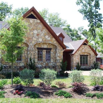 Single-story stone house with gabled roof, landscaped front yard, and flower beds on a sunny day.