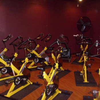 A group of yellow stationary exercise bikes arranged in a fitness studio with dark red walls and a large fan.