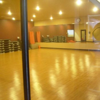 Empty fitness studio with wooden floors, mirrors, stacked step platforms, and exercise mats, viewed through a glass window.