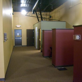 Empty office hallway with maroon and green cubicle dividers, beige walls, brown carpet, and exposed ceiling pipes.