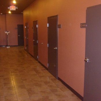 A hallway with brown doors on both sides, tan tiled floor, and a weighing scale at the far end near a closed door.