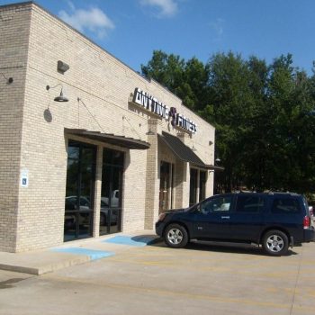 A dark SUV parked in front of a brick Anytime Fitness gym building with accessible parking and trees in the background.