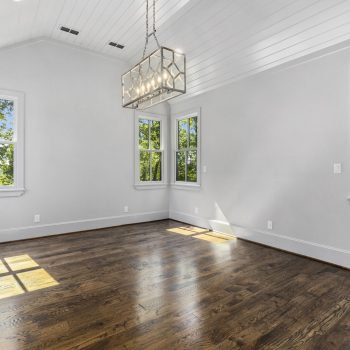 Empty room with large windows, white walls, wood floors, and a modern rectangular chandelier hanging from the ceiling.