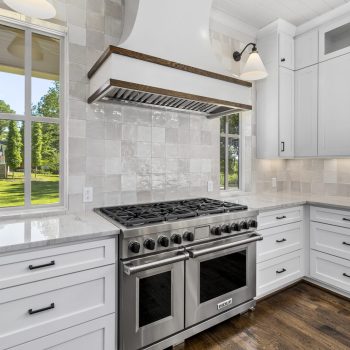 Modern kitchen with white cabinets, stainless steel gas stove, large hood, tile backsplash, and two windows with outdoor view.