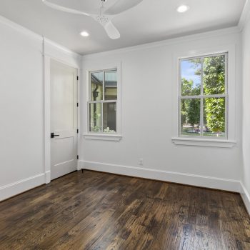 Empty room with white walls, two windows, a ceiling fan, and hardwood flooring. Daylight comes through the windows.