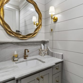 Small modern bathroom with gray vanity, marble countertop, gold fixtures, round mirror, and white shiplap walls.
