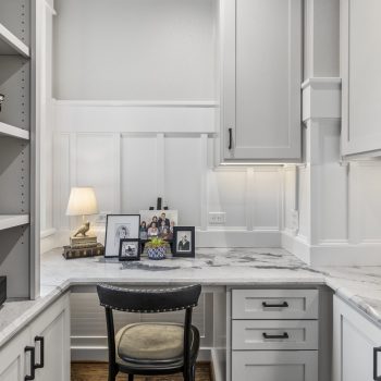 A home office corner with white cabinetry, marble countertop, a chair, family photos, books, and a desk lamp.