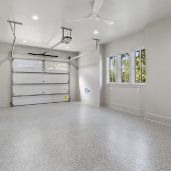 Empty modern garage with speckled flooring, overhead door, ceiling fan, windows, and wall outlets.