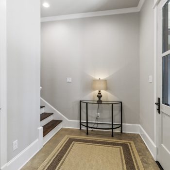 A small entryway with a rug, a glass console table, a lamp, stairs, and a door with glass panels.