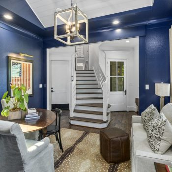 A small sitting room with blue walls, a sofa, chairs, a round table, and a staircase leading to the upper floor.