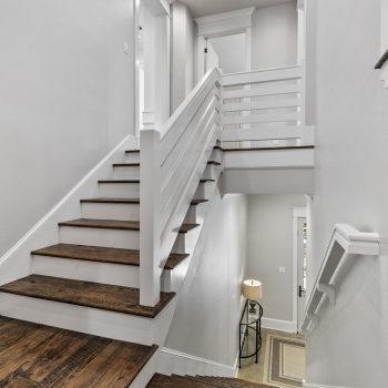 Wooden staircase with white railings in a modern, bright interior, leading to a landing and entryway with a lamp.
