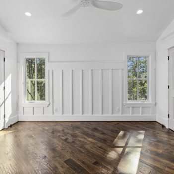 Bright empty room with white walls, two windows, double doors, a ceiling fan, and dark hardwood floor.