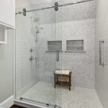 Spacious walk-in shower with glass doors, mosaic tile walls, dual shower heads, and a wooden bench with folded towels.