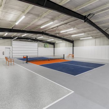 Indoor pickleball court with blue and orange markings, high ceiling, benches, ball stacks, and bright overhead lights.