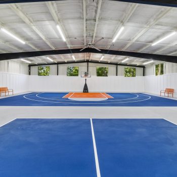 Indoor basketball court with blue and orange markings, two benches on each side, and white paneled walls.