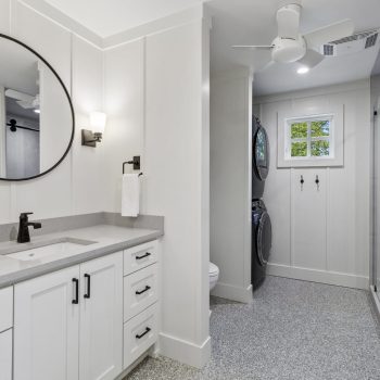 Modern white bathroom with a round mirror, vanity, stacked washer and dryer, walk-in shower, and ceiling fan.