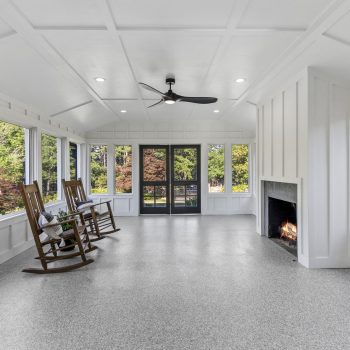 Bright sunroom with large windows, a ceiling fan, two rocking chairs, and a lit fireplace overlooking a green yard.