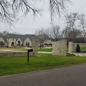 Large stone house with black roof, circular driveway, stone fence, and mailbox, set on a spacious green lawn.
