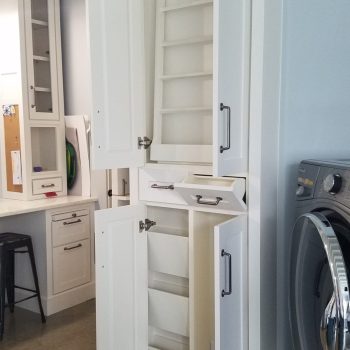 Tall white cabinet with open doors and pull-out trays, showing empty shelves beside a washer and a desk area.