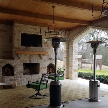 Covered patio with stone fireplace, mounted TV, patio heaters, two chairs, and view of yard with trees and playset.