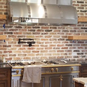 A stainless steel range with gold accents sits against a brick wall with a matching metal range hood above it.