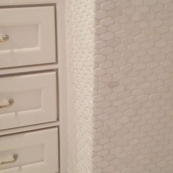 White bathroom cabinets with gold handles next to a wall covered in white fish scale tile with marble baseboards.