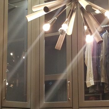 Modern chandelier with exposed bulbs hangs above an organized closet with glass-front cabinets and hanging clothes.