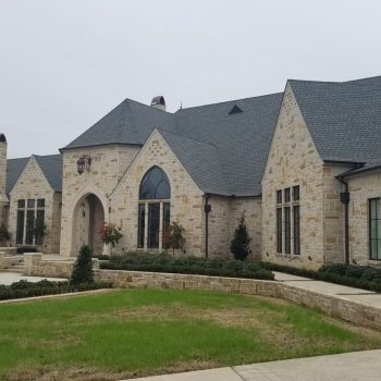 Large stone house with steep gray roof, arched entryway, tall windows, and landscaped front yard on a cloudy day.
