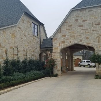 A large stone house with an arched carport, manicured shrubs, and a white SUV parked in the background.