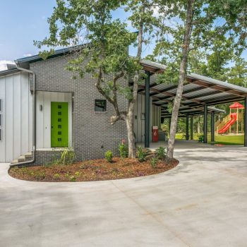 Modern house with gray brick and metal siding, green door, covered carport, and a red slide in a green yard.