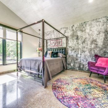Bedroom with a metal canopy bed, purple chair, colorful rug, large window, and textured accent wall.